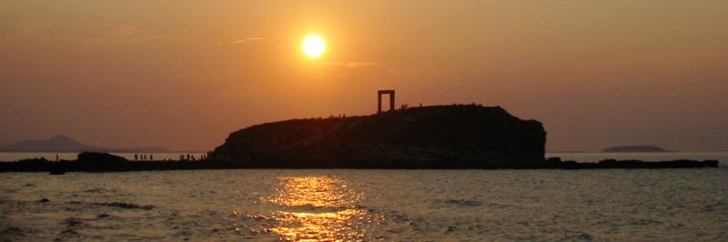Panoramic sunset view of Portara Gateway on Naxos, Greece. The ancient marble doorway stands silhouetted atop a rocky peninsula surrounded by the Aegean Sea. Golden sunlight reflects off rippling waters, with distant islands visible on the horizon. Small figures of tourists can be seen against the dusky sky.